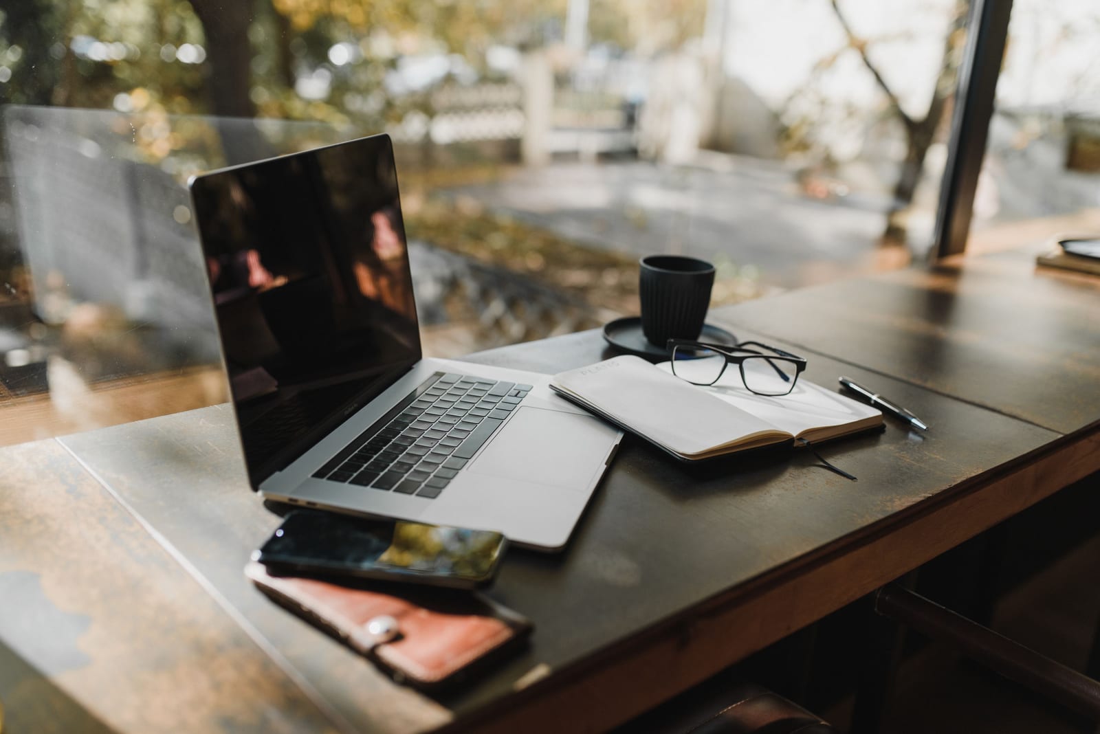 A laptop, notebook, glasses and coffee on a wooden desk by a window
