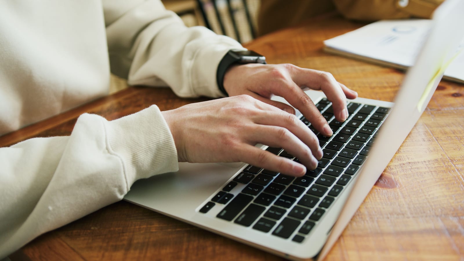 Hands typing at a wooden desk with a document beside the laptop