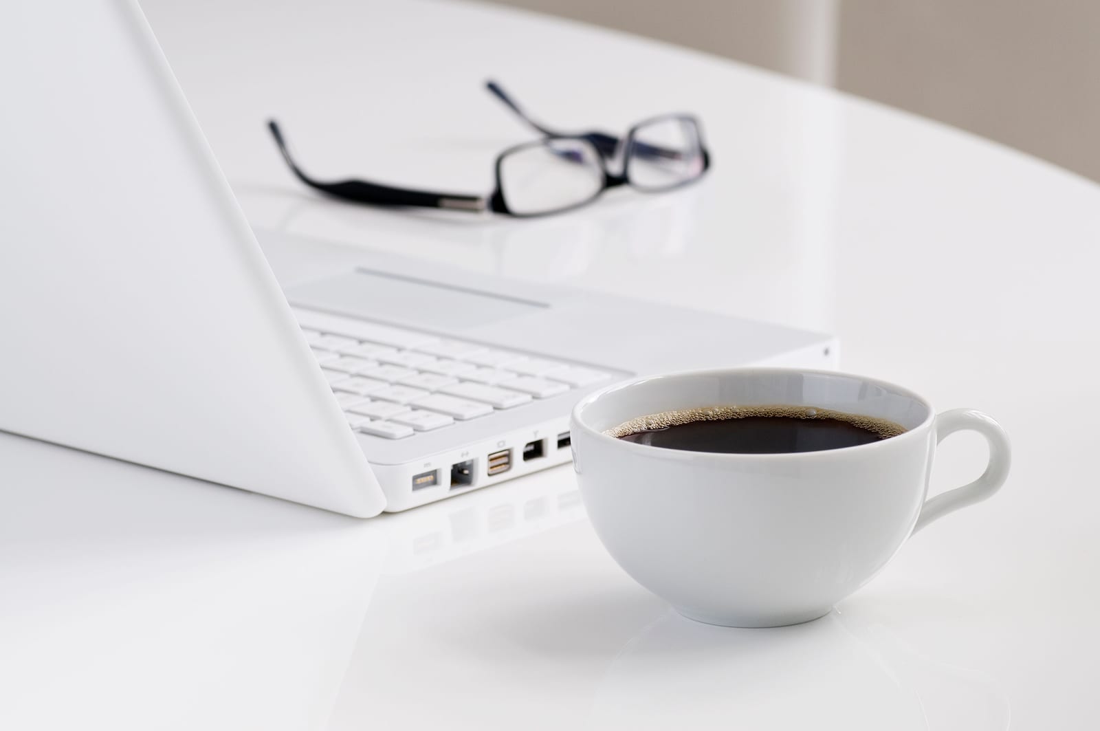 A white laptop, a cup of black coffee and reading glasses on a white table