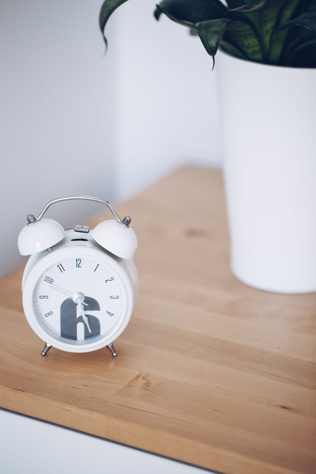 A small white twin-bell alarm clock on a wood desk beside a planter