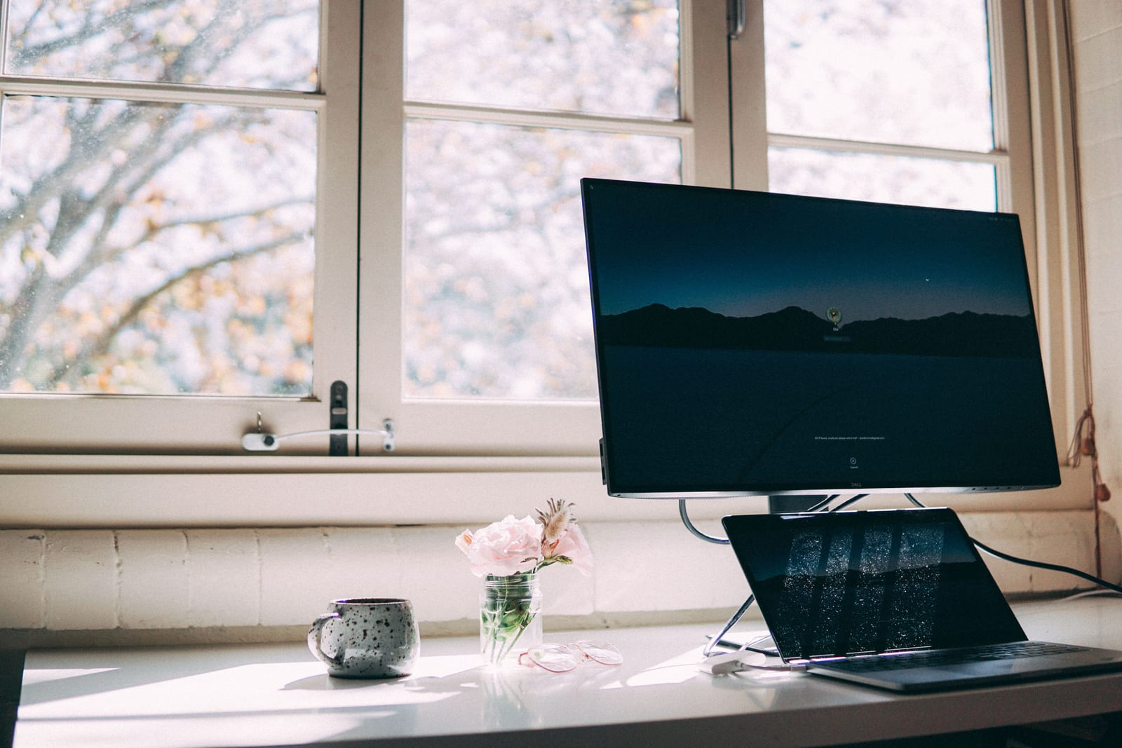 A home-office desk by a window with a laptop, mug and flowers in soft daylight