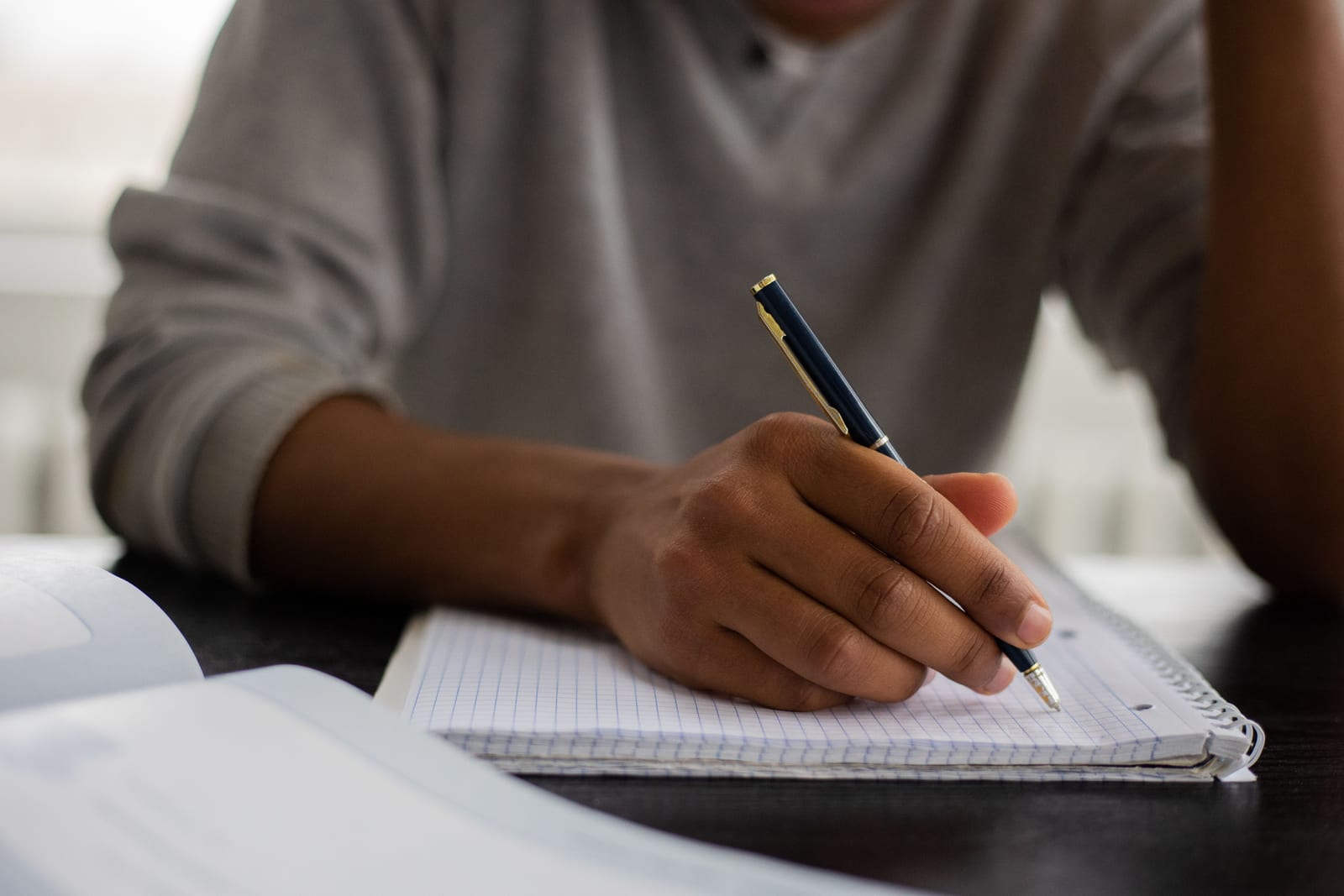 A hand writing notes in a grid notebook beside an open book