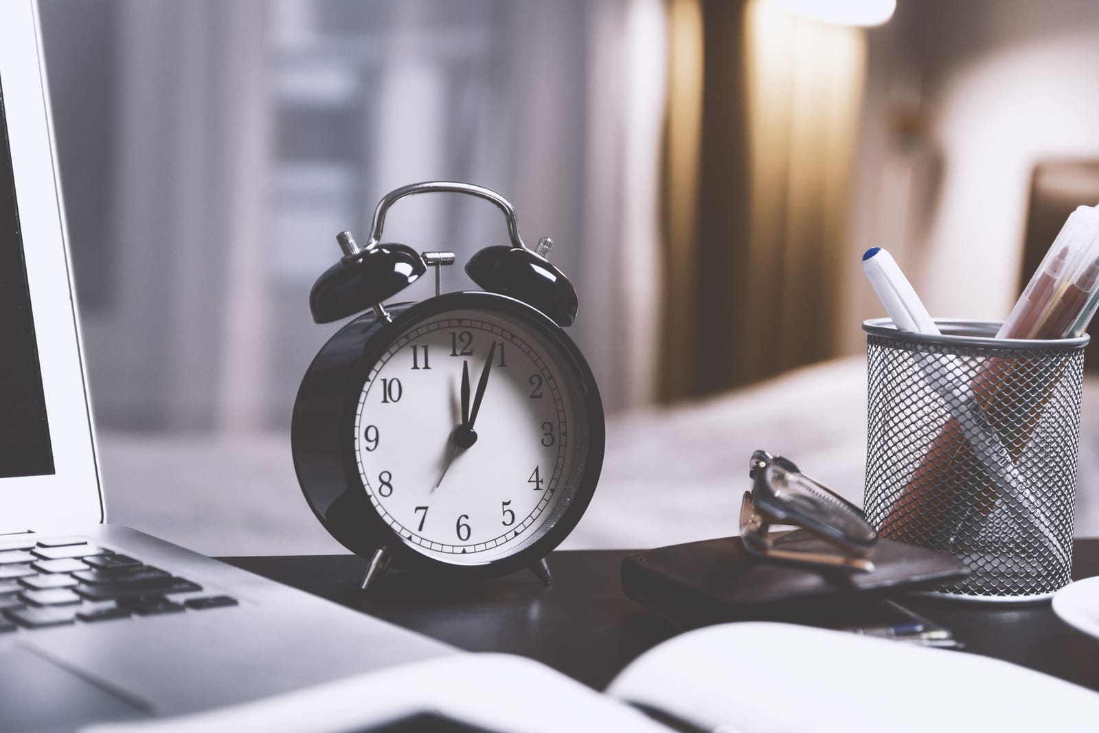 An alarm clock on a desk beside the edge of a laptop and a pen holder