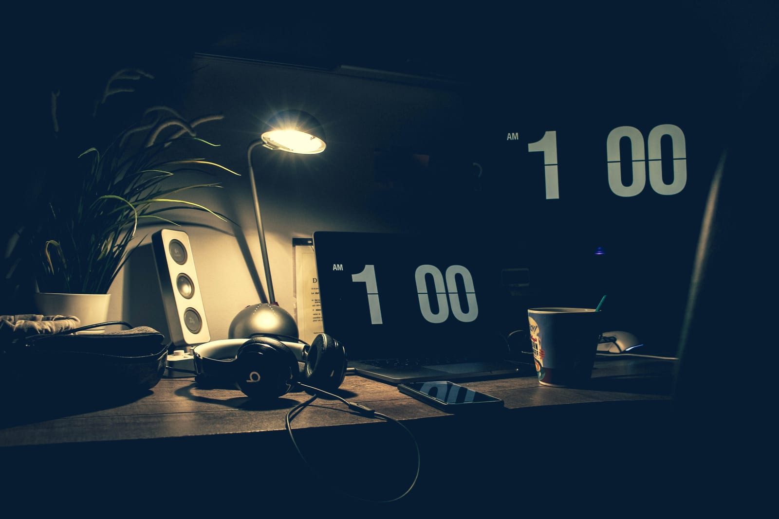 A dark home desk lit by a warm lamp with a laptop and a flip clock reading past one