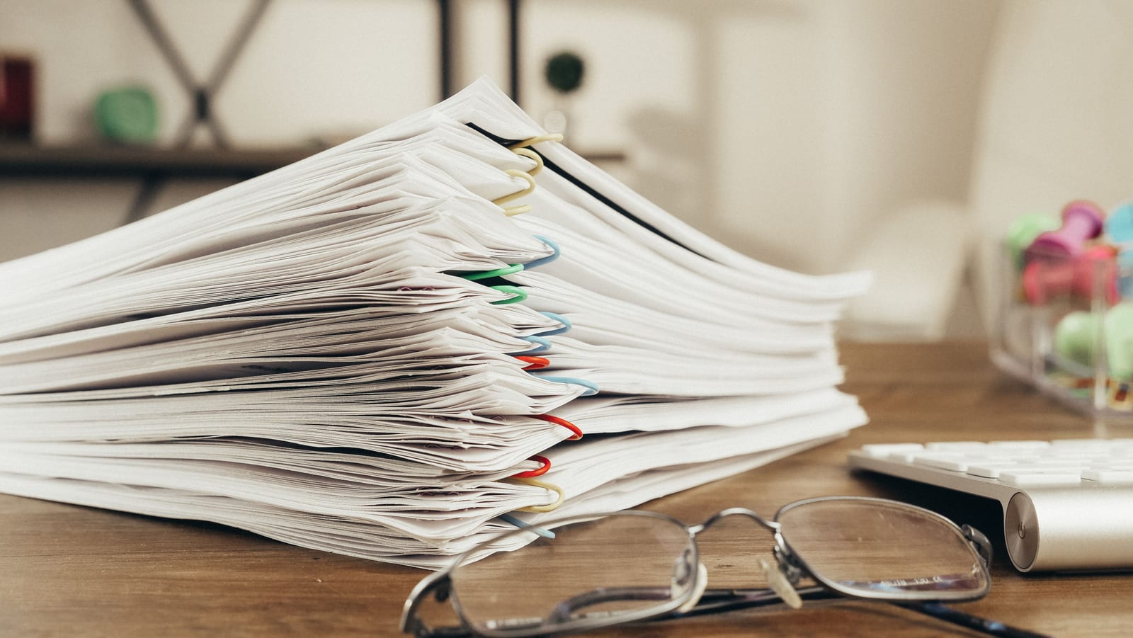 A towering stack of clipped paper documents beside glasses and a keyboard