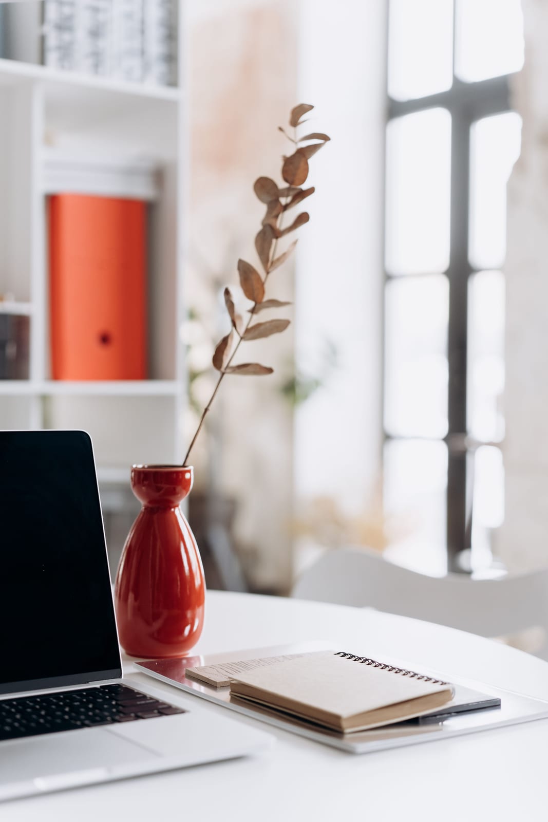 An open laptop on a white desk beside a notepad and a vase with a dried branch