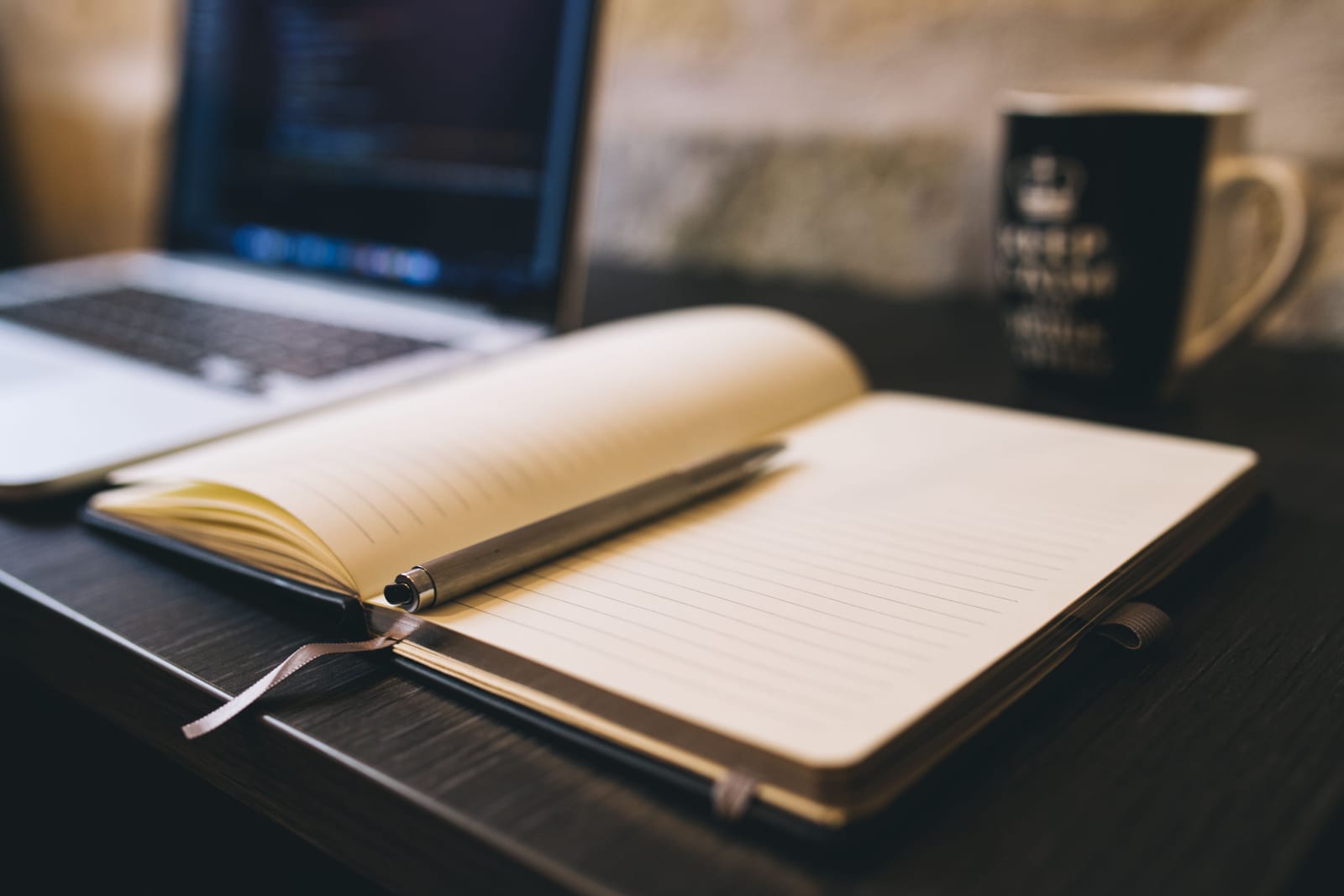 An open blank notebook and pen beside a laptop on a desk