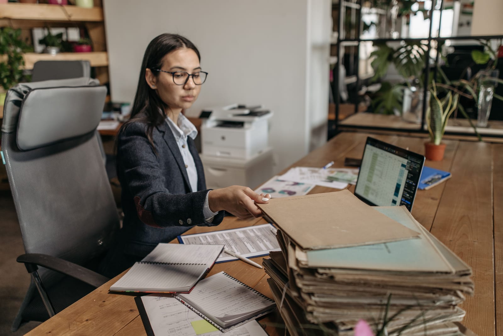 A professional at a desk beside a tall stack of paper files and a laptop