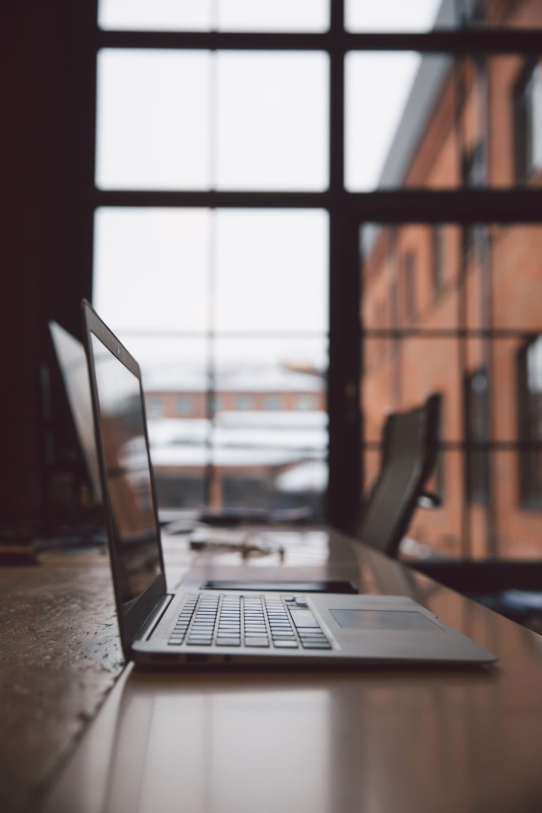 An open laptop on a wooden desk by a tall industrial window